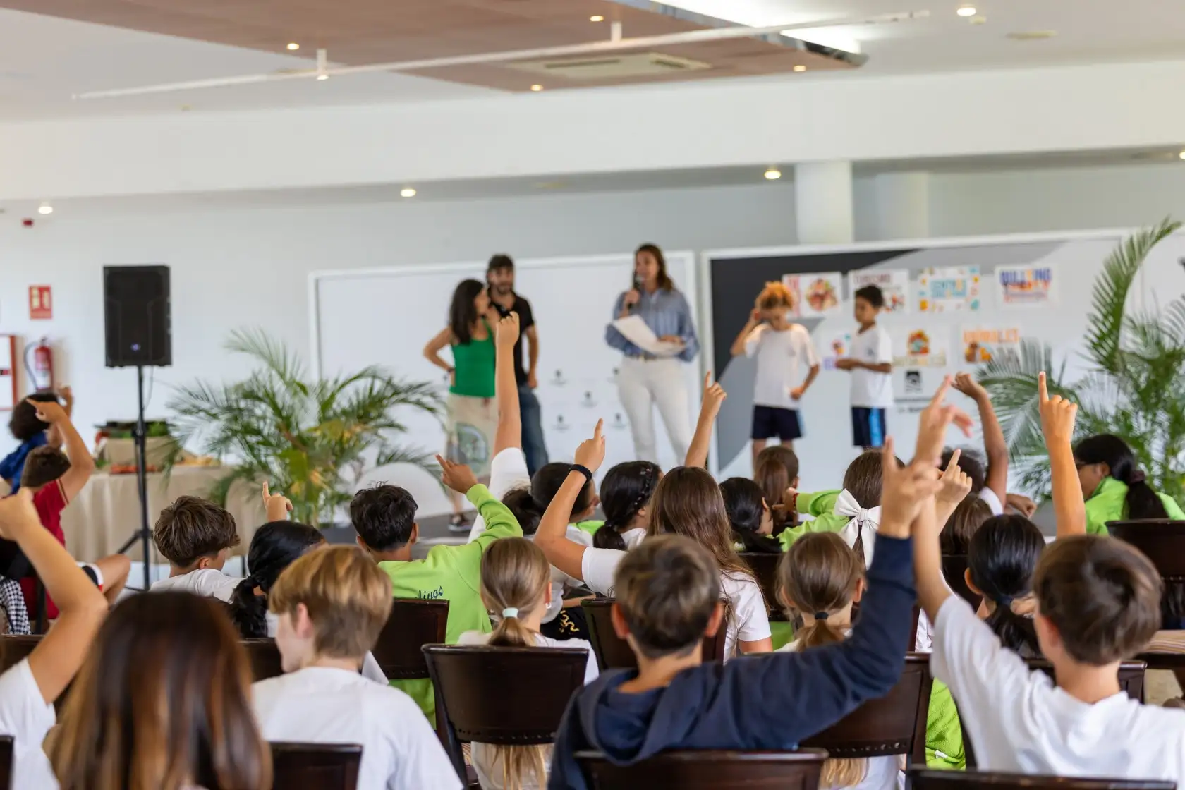 Salón lleno de niños y niñas con las manos levantadas para participar en la sesión de clausura junto a la concejal del Área Raquel Rodríguez